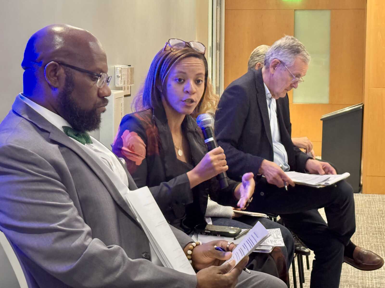 Three panelists seated indoors: a man in a gray suit and green bow tie, a woman speaking into a microphone, and another man in a blazer reviewing papers.