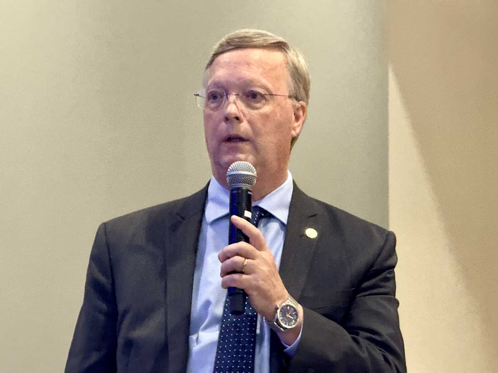Man in a dark suit and blue dotted tie speaks into a handheld microphone.