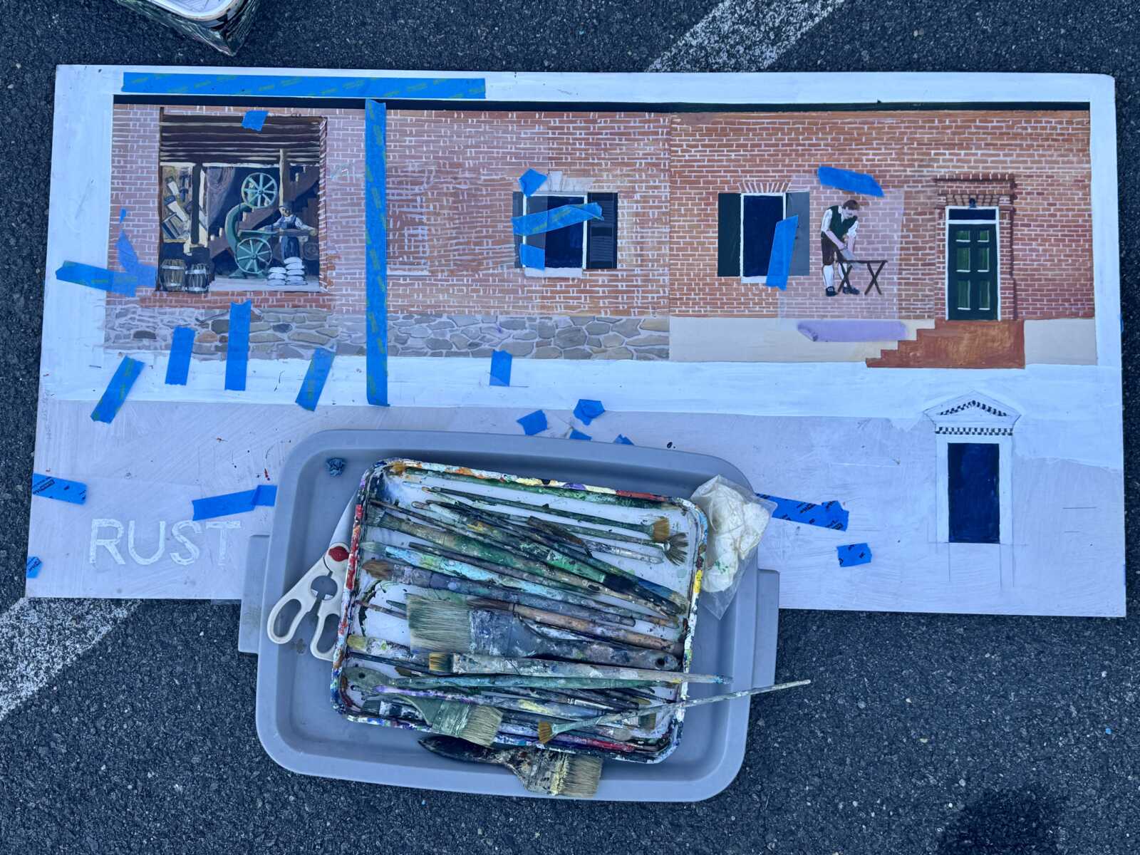 Outdoor art project: a large mural of a brick building taped to the ground with blue painter's tape; painter's tray and brushes in the foreground.