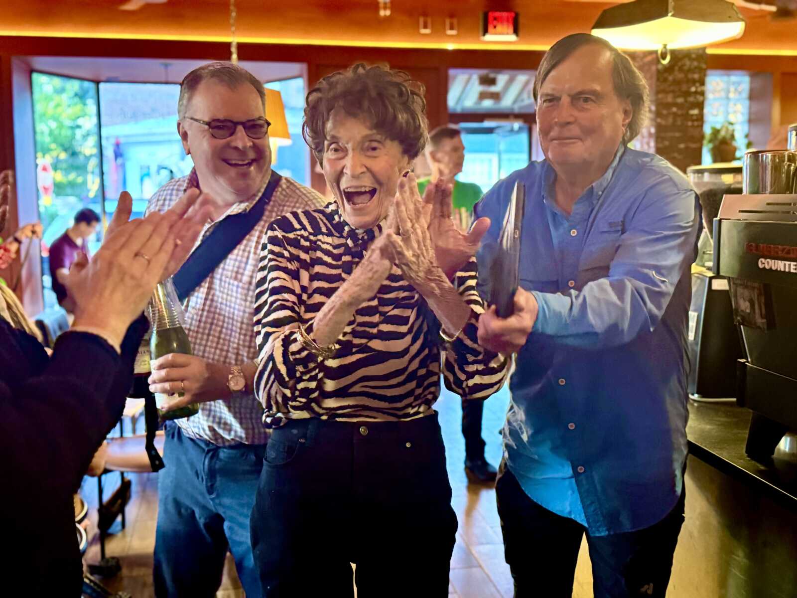 Three adults cheer and clap inside a cafe, woman in zebra-print top at center smiling widely, man on her left holding a bottle, man on her right in blue shirt joins the celebration.