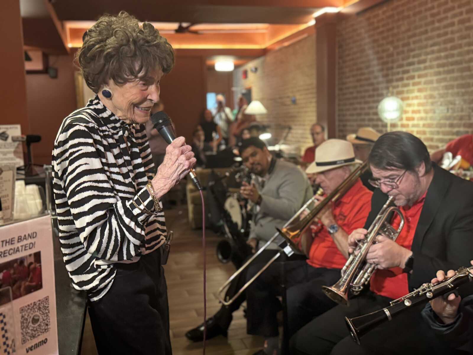 Elderly woman singing into a microphone while a brass band plays in a brick-walled venue.