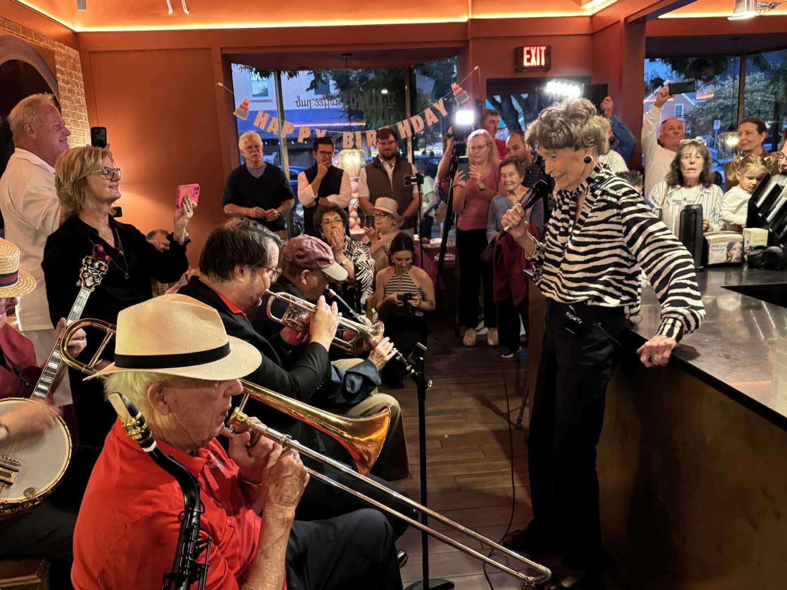 Older woman in a zebra-print blouse sings into a microphone as a brass band performs for a birthday party.