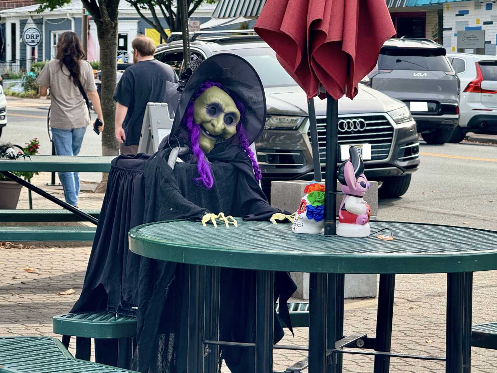 IMG_6302 morning | ALXnow Life-size witch figure in a black cloak with purple braids sits at a round green outdoor table on a busy street.