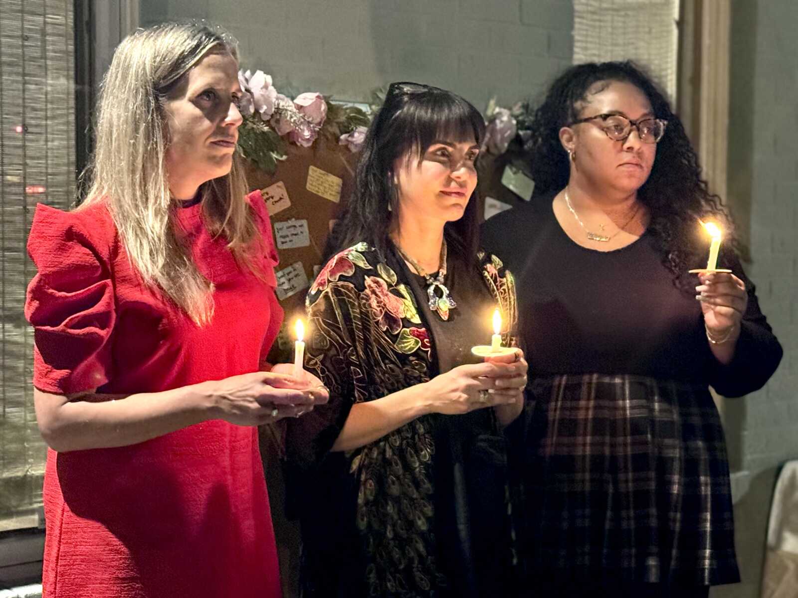 Three women stand in a candlelight vigil, each holding a lit candle and looking thoughtful.
