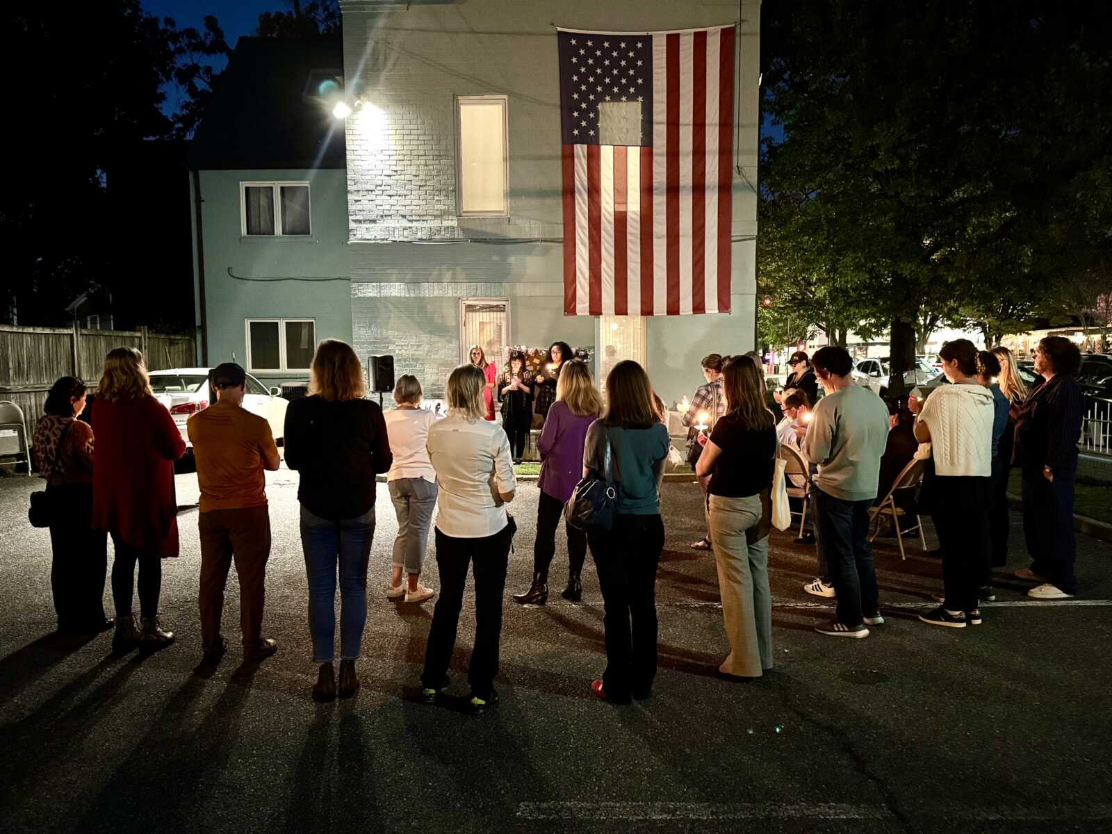 Group of people gathers outside a building at night for a candlelight vigil beneath a large American flag banner outside.
