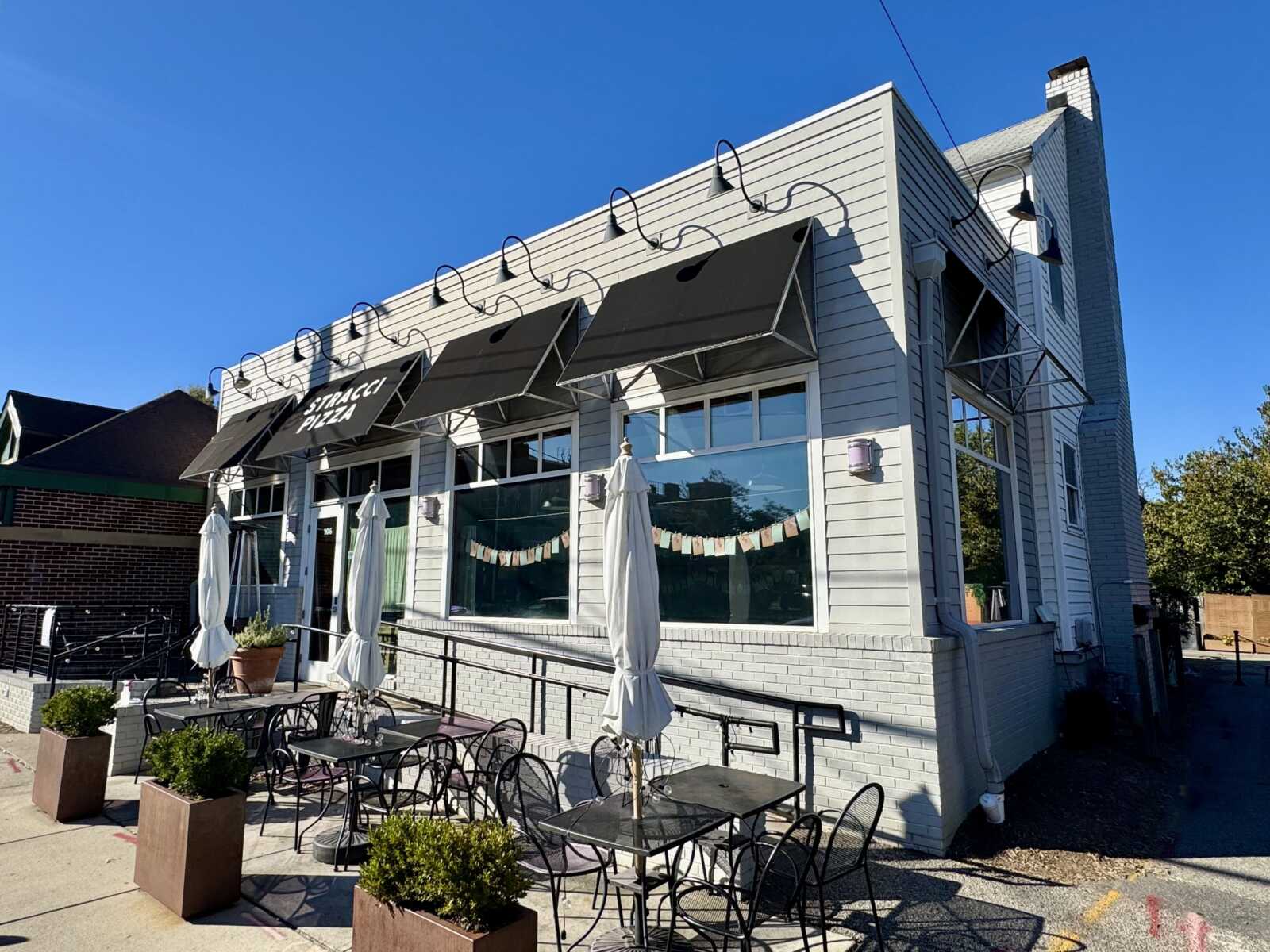 Stracci Pizza in Del Ray (staff photo by James Cullum) | ALXnow White brick cafe with multiple black awnings, outdoor tables and chairs, and potted plants in the foreground.