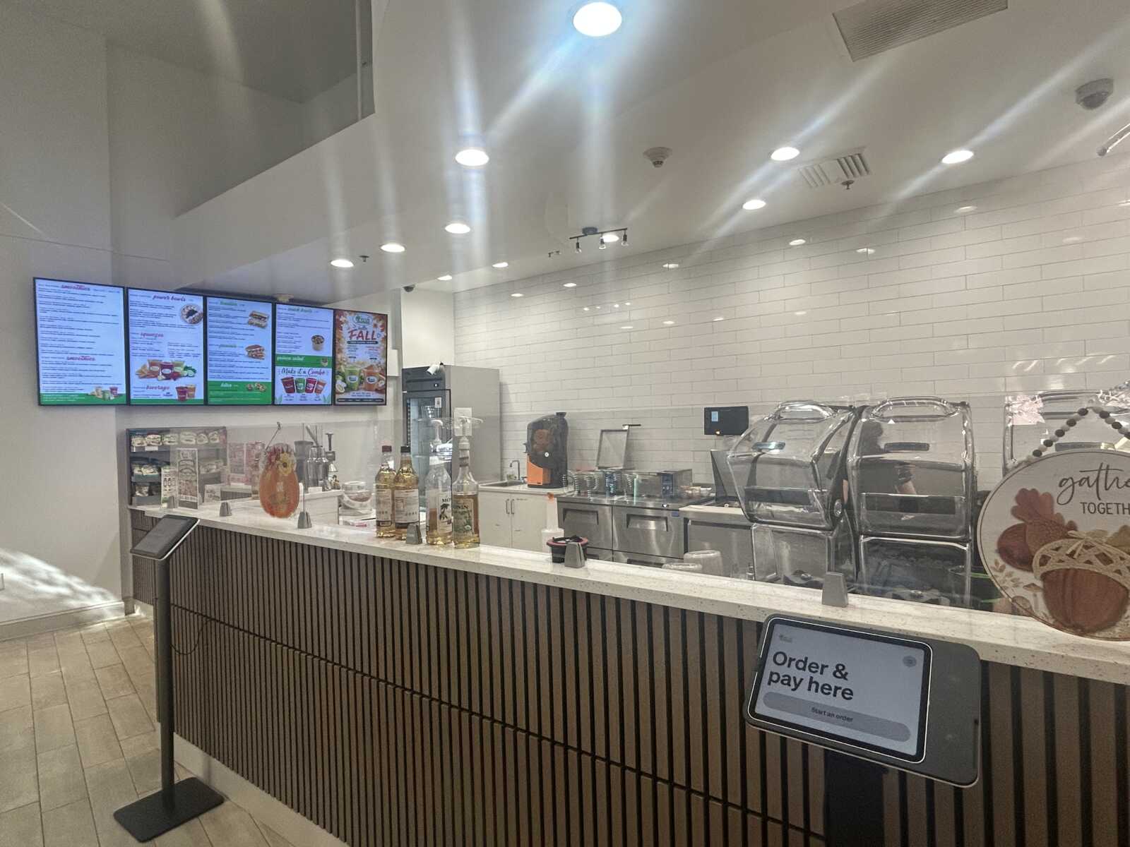 Cafe counter with digital menu boards on the wall and a self-serve order/pay kiosk at the front.