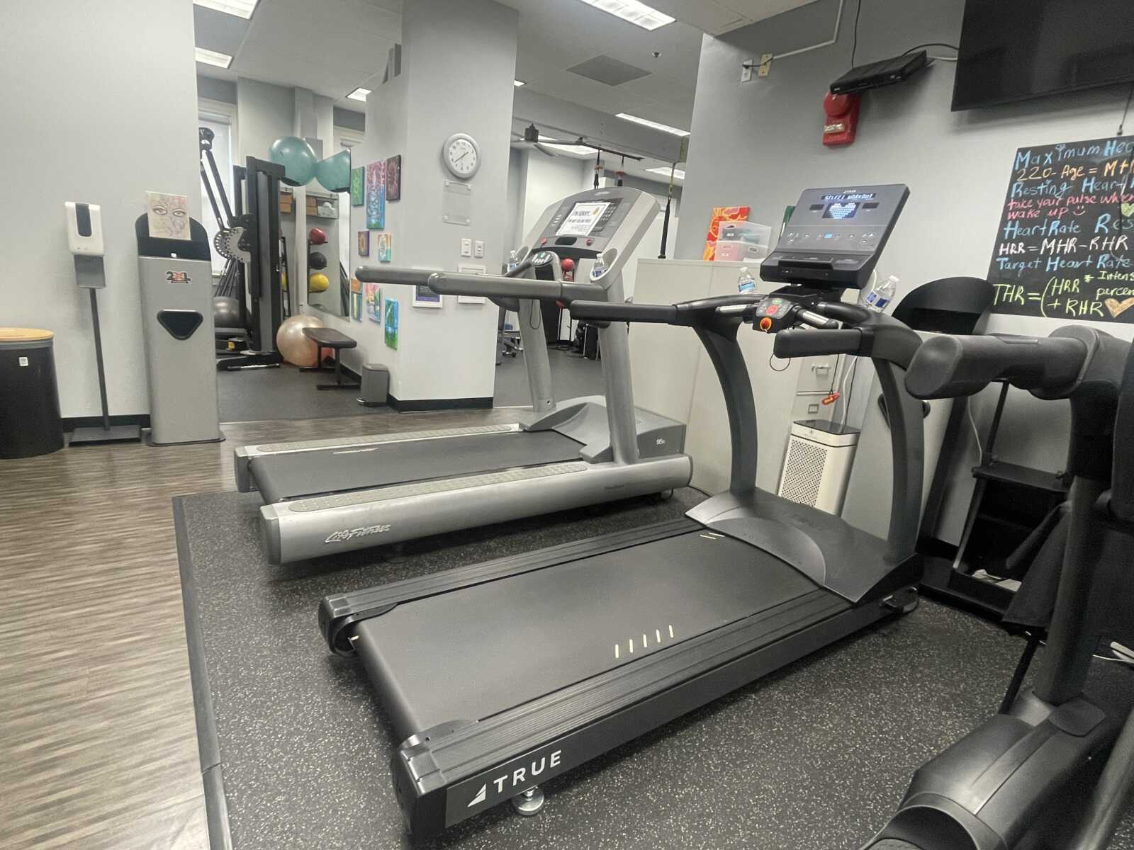 Two treadmills in a modern gym with other cardio machines and a chalkboard in the background.