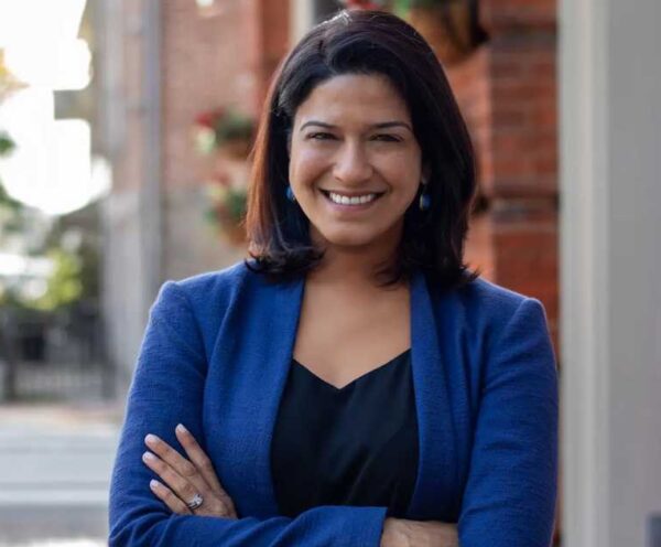 Smiling woman with shoulder-length dark hair wearing a blue blazer and black top, standing with arms crossed outdoors.