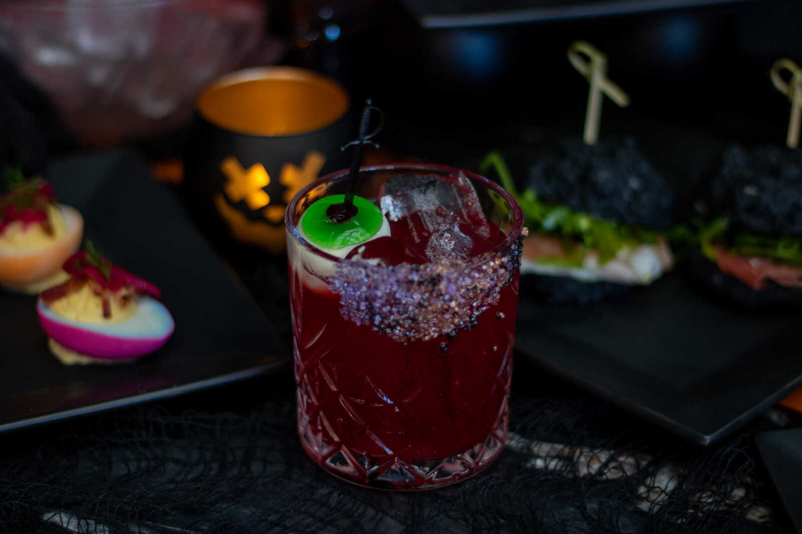 A ruby red cocktail in a patterned glass with a sugared rim, ice cubes, and a green lime garnish on a dark bar or table.