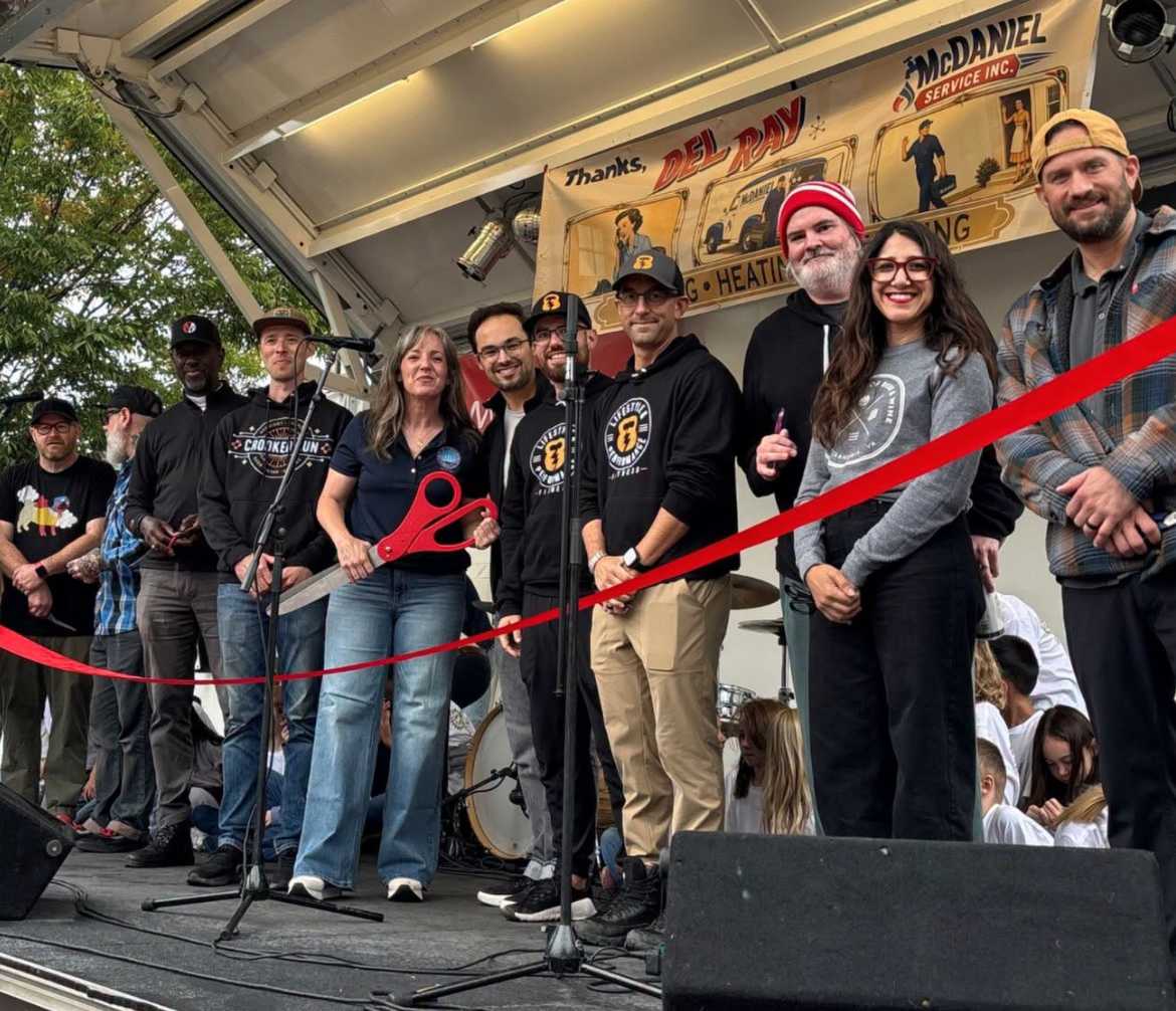 Group of people on a stage during a ribbon-cutting ceremony, a woman holding oversized red scissors is prominent in the foreground.