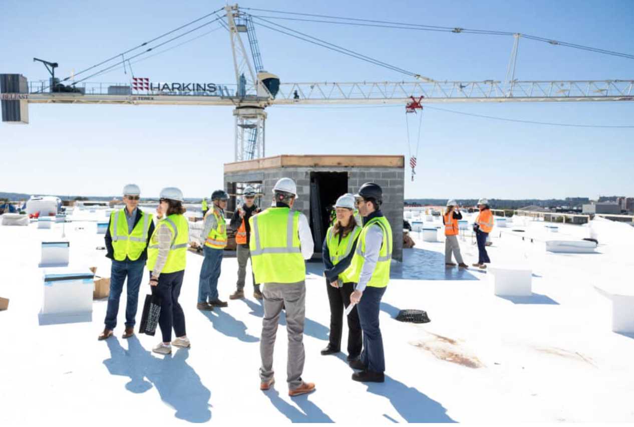 A group of construction workers and supervisors in hard hats and high-visibility vests stand on a white rooftop next to a small brick structure, with a crane in the background.