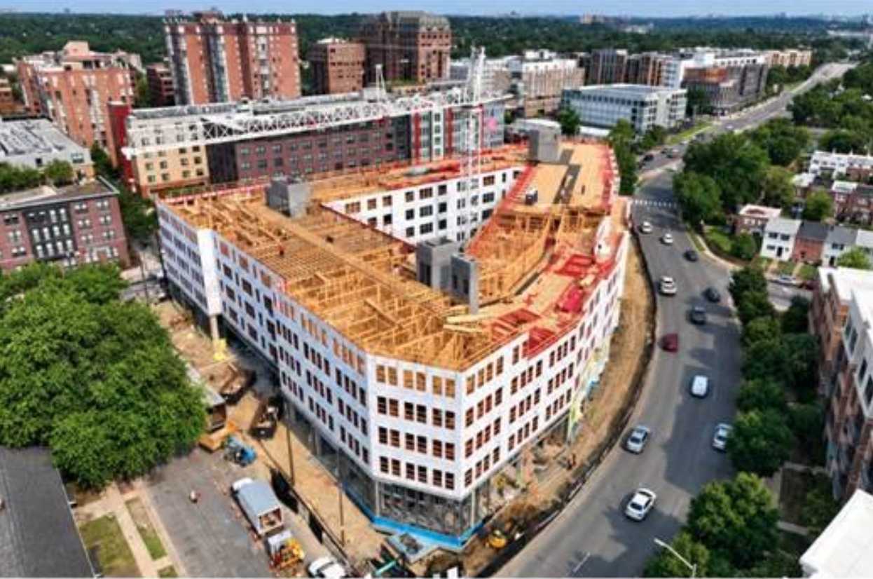 Aerial view of a curved, white, multi-story building under construction in a downtown block, with orange scaffolding and surrounding streets.