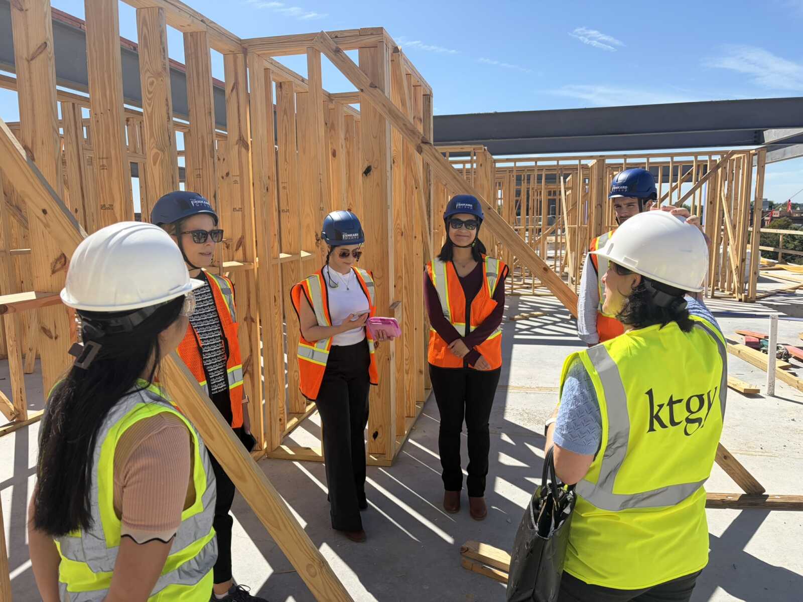 Group of construction workers in hard hats and high-visibility vests standing in a wooden-frame building under construction on a sunny day.