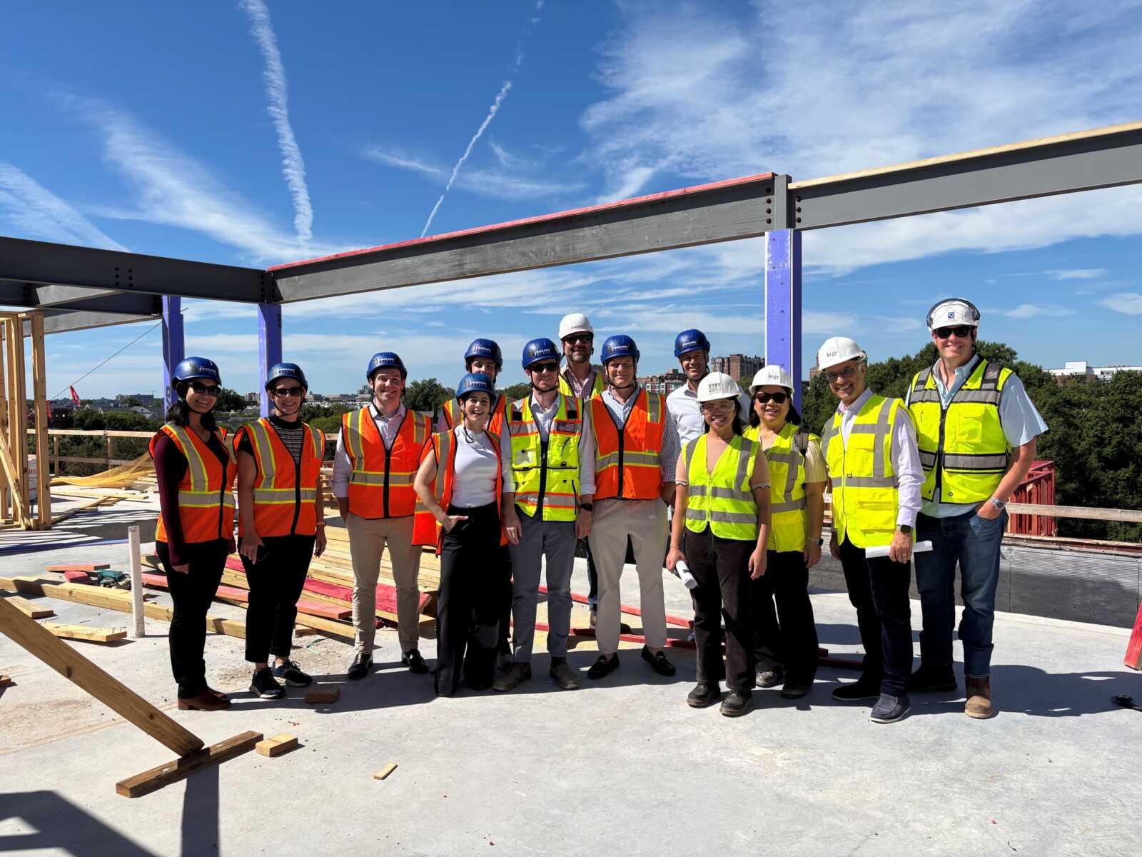 Group of construction workers posing on an unfinished rooftop, wearing hard hats and high-visibility vests under a blue sky.