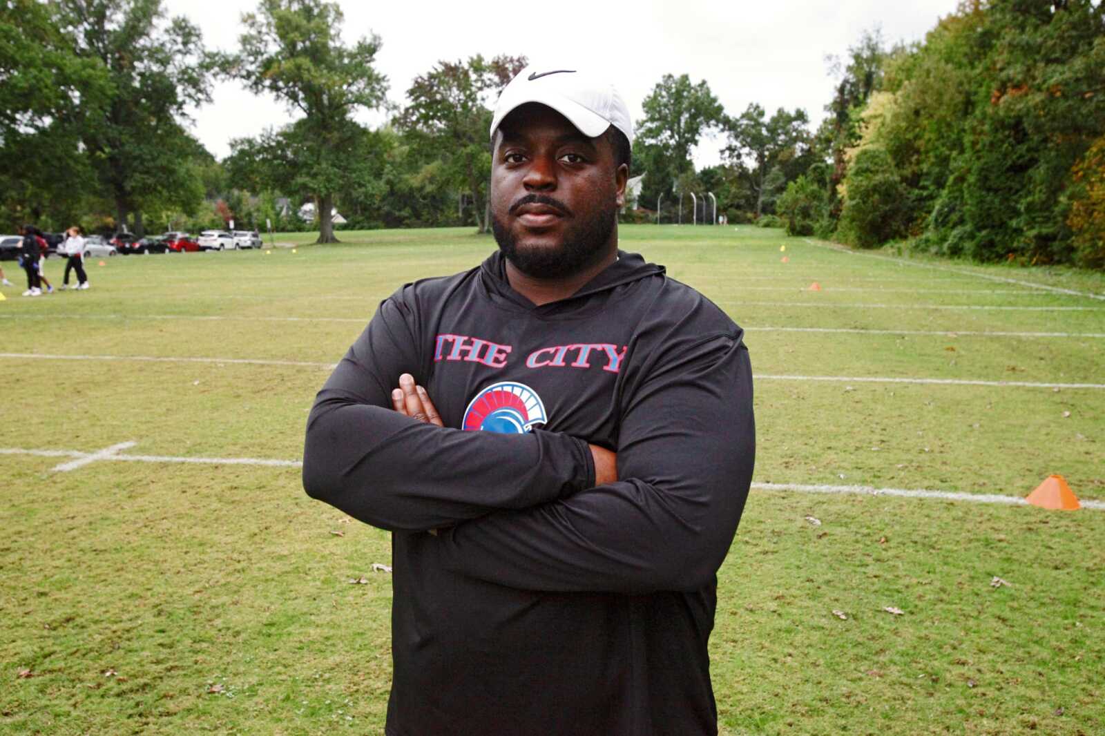 Man in a black hoodie with 'THE CITY' logo and a white cap stands with arms crossed on a grassy sports field.