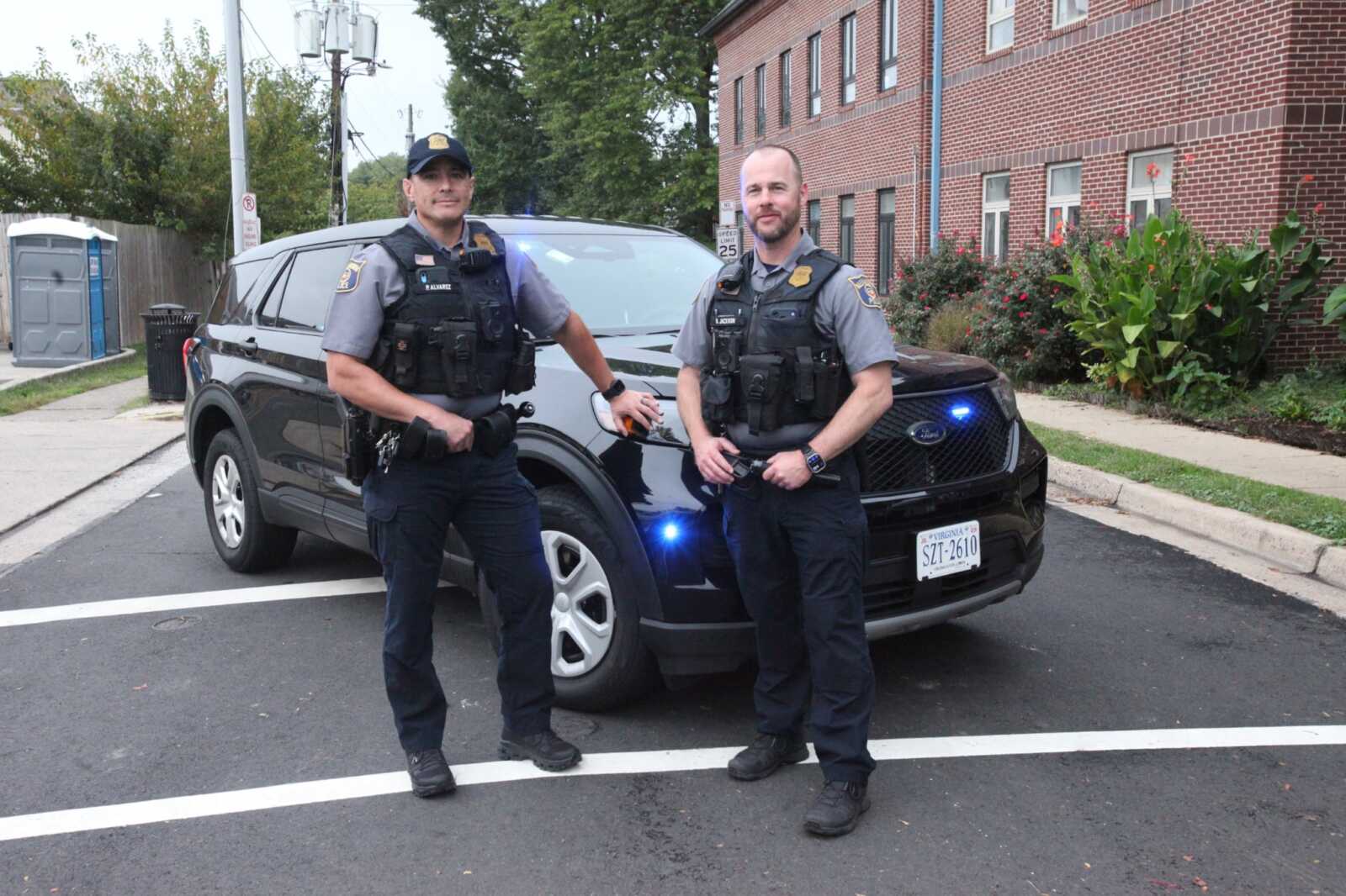 Two police officers stand beside a dark police SUV with blue lights in a residential street in front of a brick building and sidewalk