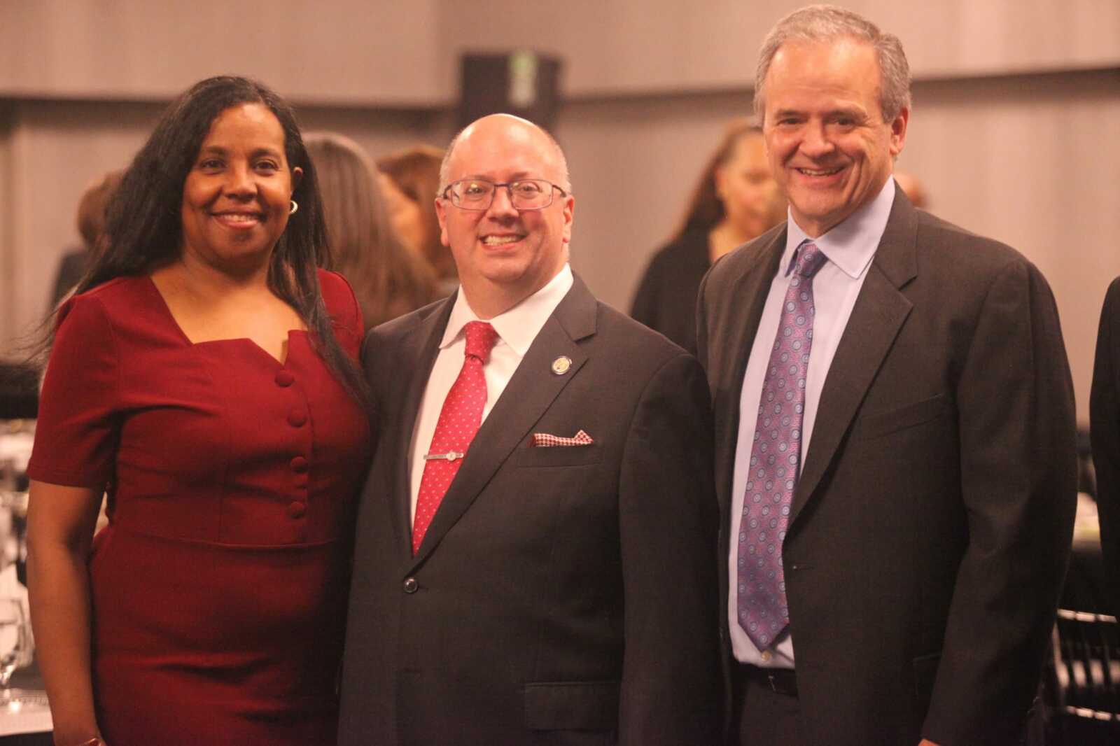 Three smiling adults in formal attire posing for a photo at a banquet: a woman in a red dress on the left, and two men in suits and ties in the center and right.