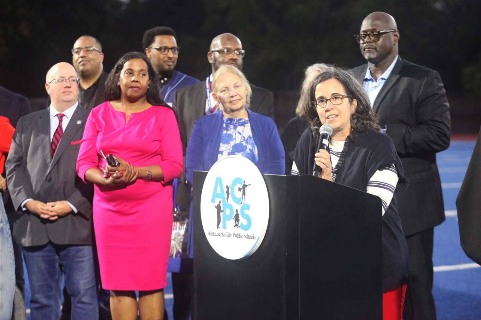 Woman with glasses speaks into a microphone at a podium with the ACPS logo, as diverse officials stand behind her on stage.
