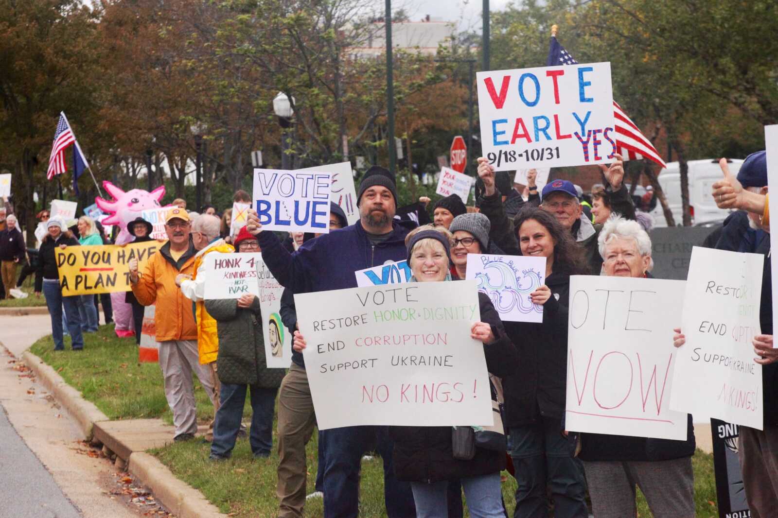 Crowd of protesters holding handmade signs with messages like 'VOTE EARLY' and 'NO KINGS' at a street-side demonstration.