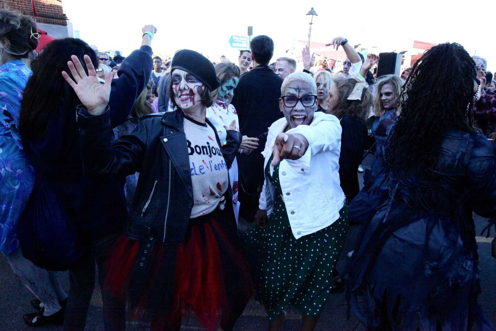 Crowd of people in Halloween costumes dancing in a street, two women in the foreground with face paint smiling and raising their hands.