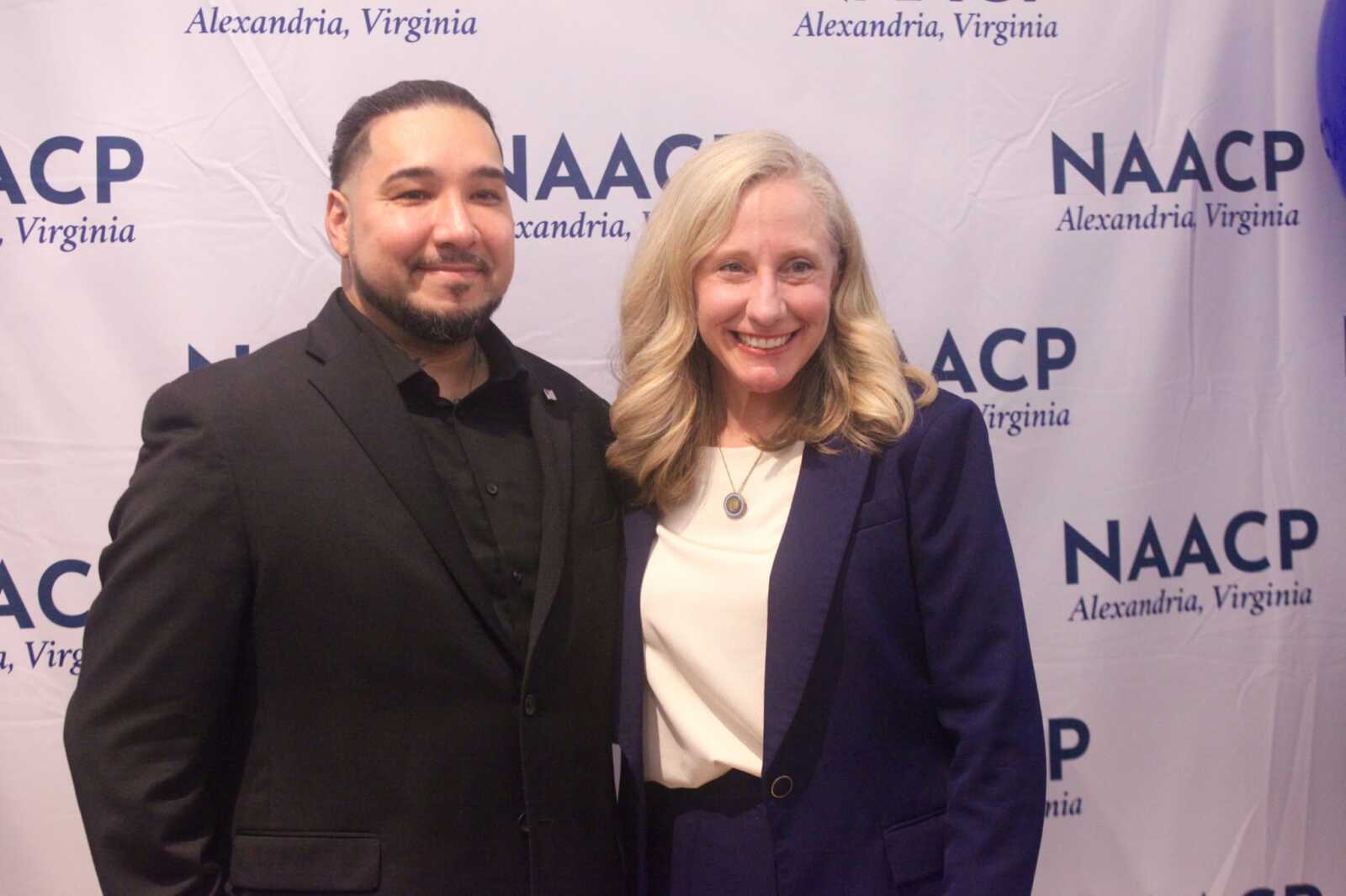Two smiling adults pose together at a NAACP event, with a man in a black suit and a woman in a navy blazer standing in front of a backdrop that reads 'NAACP Alexandria, Virginia'.