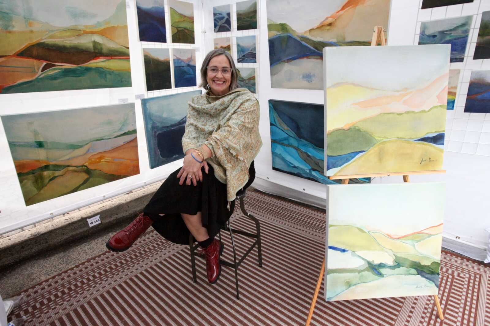 A smiling woman seated on a stool in an art gallery surrounded by colorful landscape paintings on walls and easels.