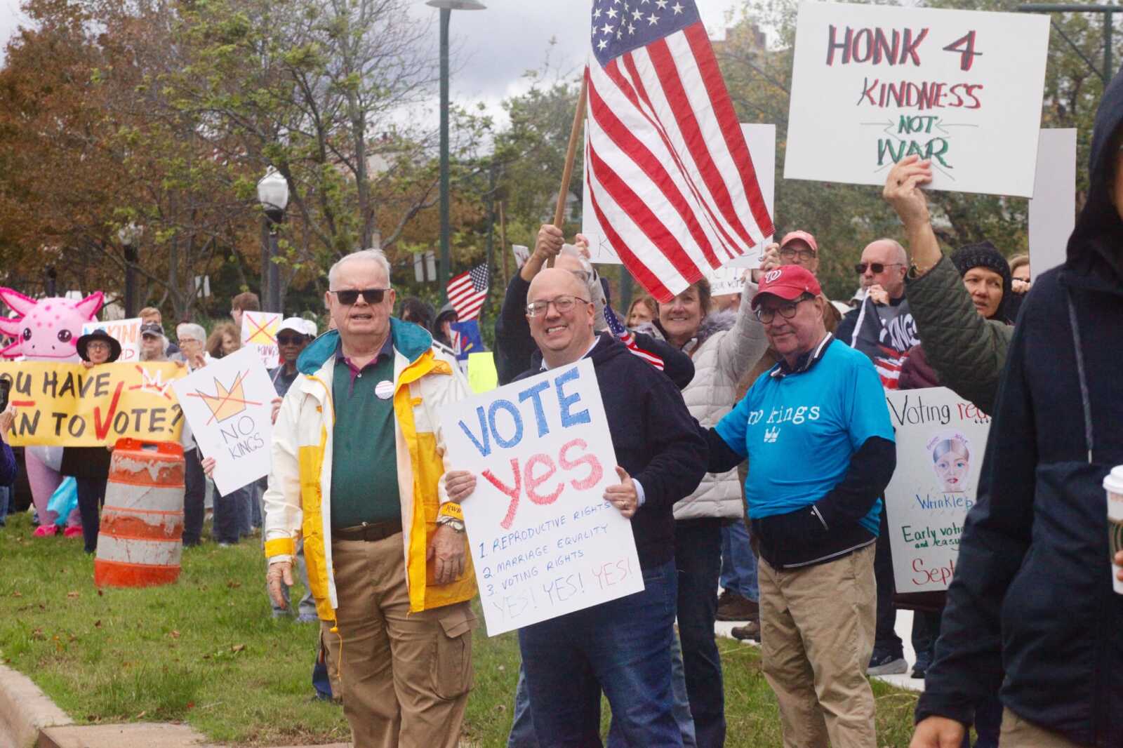 Crowd of diverse protesters holding signs like "VOTE YES" and an American flag at a rally for voting rights and kindness.