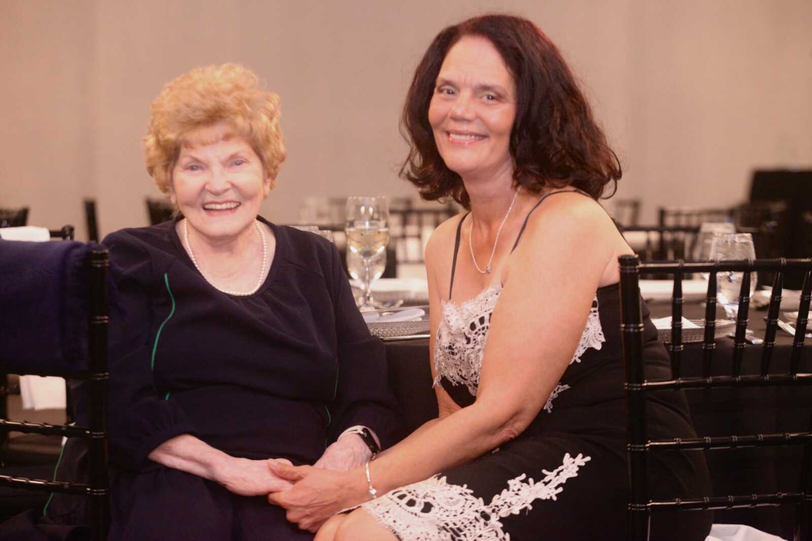 Two smiling women sit side by side at a banquet table, an older woman with short blonde hair in a dark dress and a younger woman with dark hair in a black lace dress.