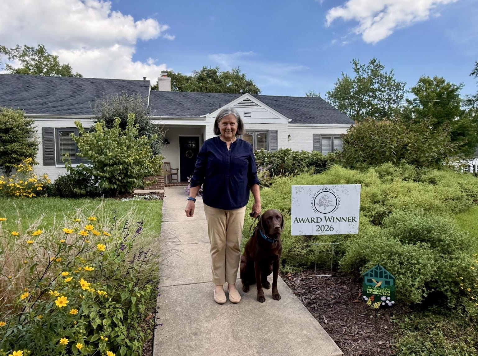 Older woman in navy blouse and tan pants standing with brown dog on a leash in a front yard, house in background and an 'Award Winner 2026' sign nearby.