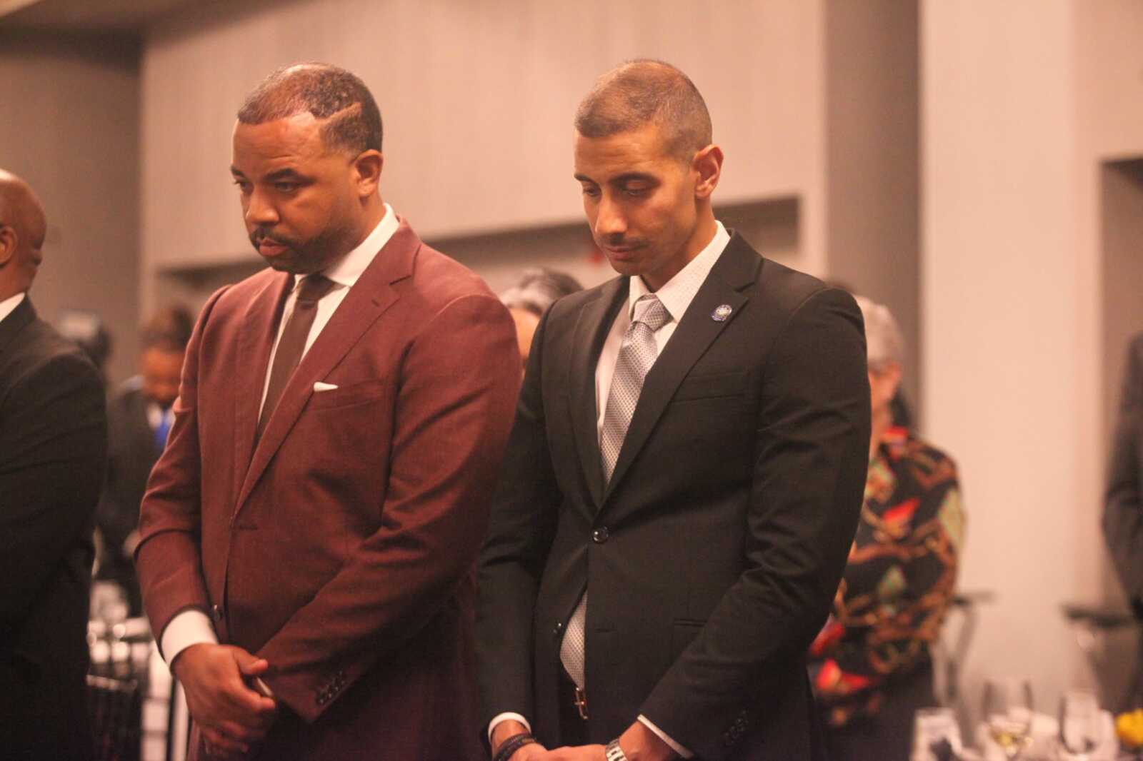 Two men in formal suits stand side by side, heads bowed at a dimly lit event hall. (Informative alt text for a formal ceremony)