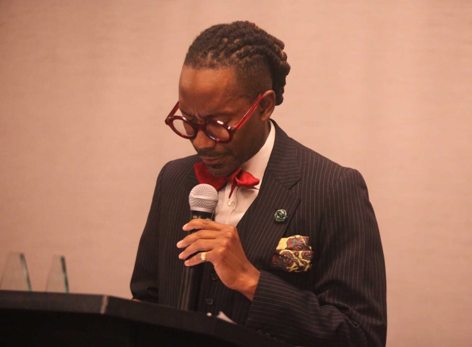 Man in a pinstripe suit with a red bow tie and glasses speaking into a handheld microphone at a podium.
