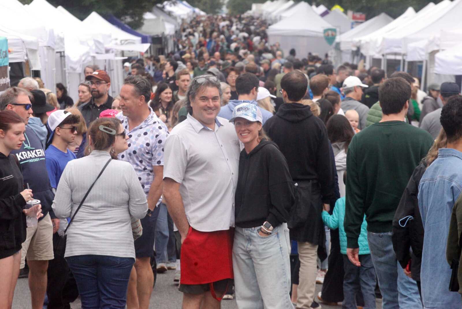 Smiling couple posing for a photo in a crowded outdoor market with white vendor tents in the background.