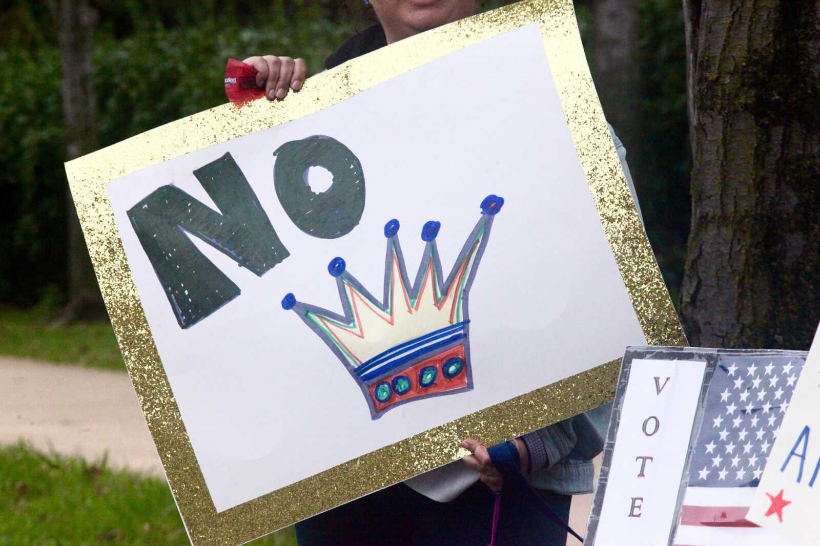 Person holding a poster with the word NO and a colorful crown drawing in a glittery gold frame outdoors.