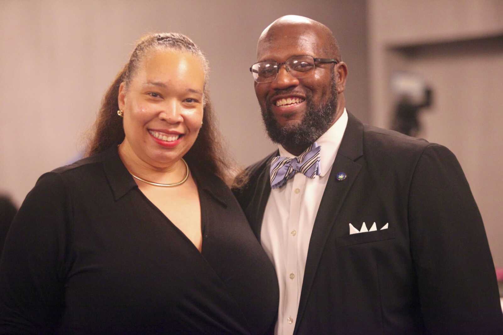 Two smiling adults pose together at a formal event; woman in a black dress with gold necklace, man in a suit and bow tie with pocket square and glasses