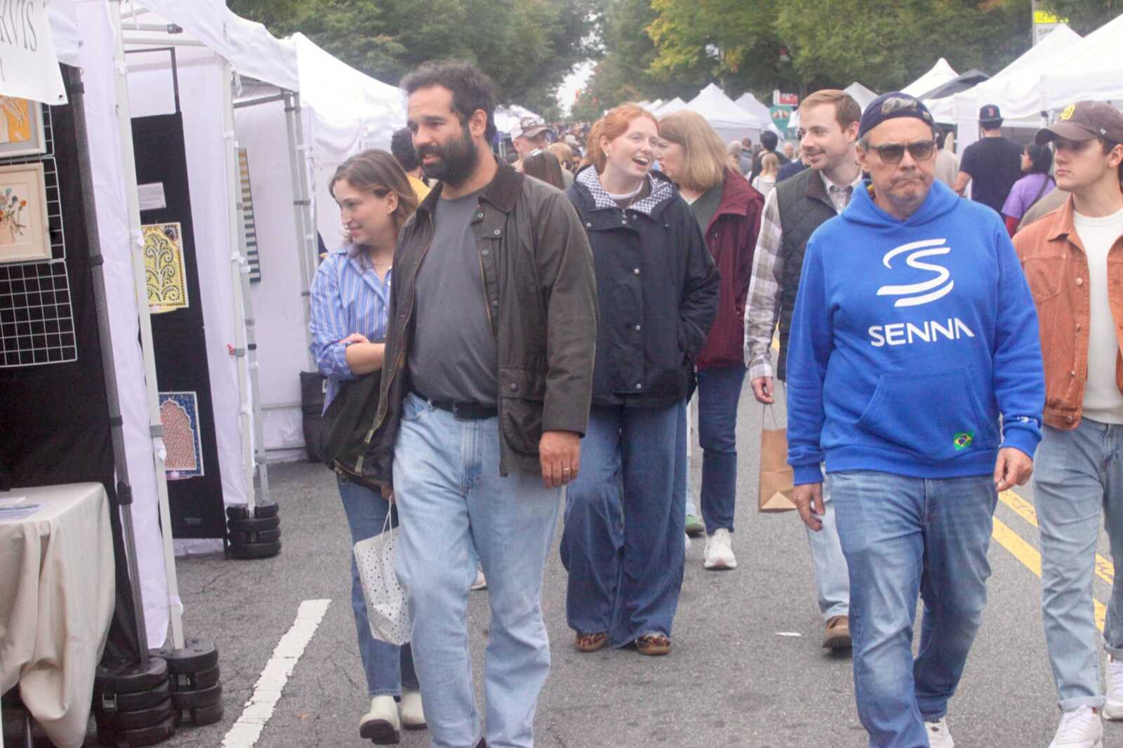 Crowd walks along an outdoor market with white vendor tents on both sides, browsing displays.