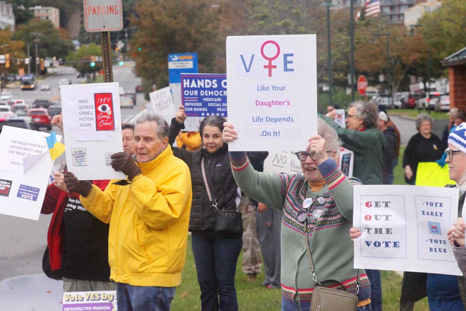 A diverse group of protesters on a city street, holding signs urging people to vote and defend democracy, including a sign that says “Get Out The Vote.”
