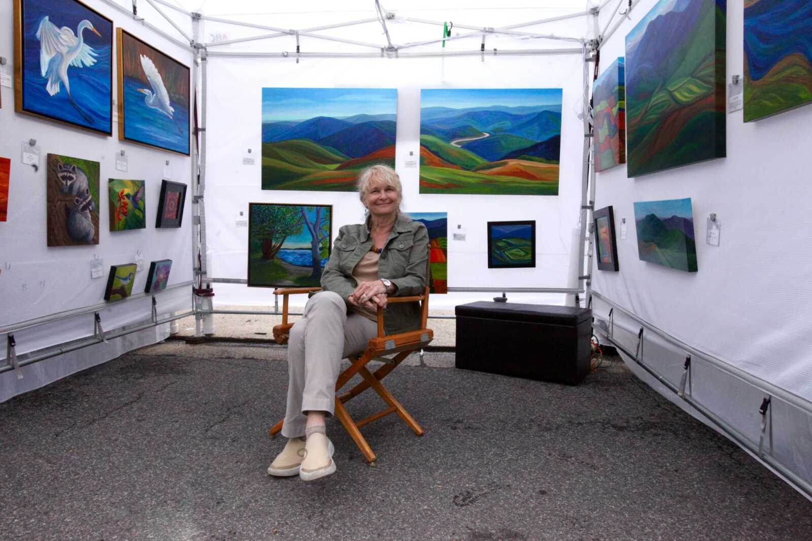 Smiling woman sits in a director's chair inside a white tent, surrounded by colorful landscape paintings on display at an art fair.