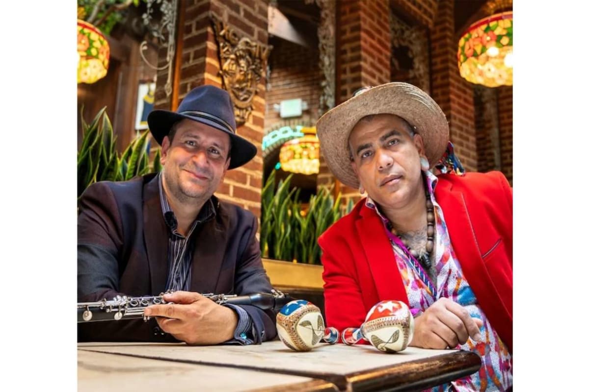 Two male musicians sit at a cafe table outdoors; one holds a flute and smiles, the other wears a straw hat and a red jacket, with decorative maracas on the table.