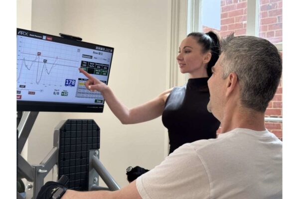 Trainer demonstrates on a cardio machine, pointing to a large monitor displaying performance metrics for a male client seated on the equipment.