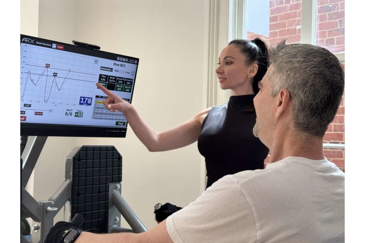 Trainer demonstrates on a cardio machine, pointing to a large monitor displaying performance metrics for a male client seated on the equipment.