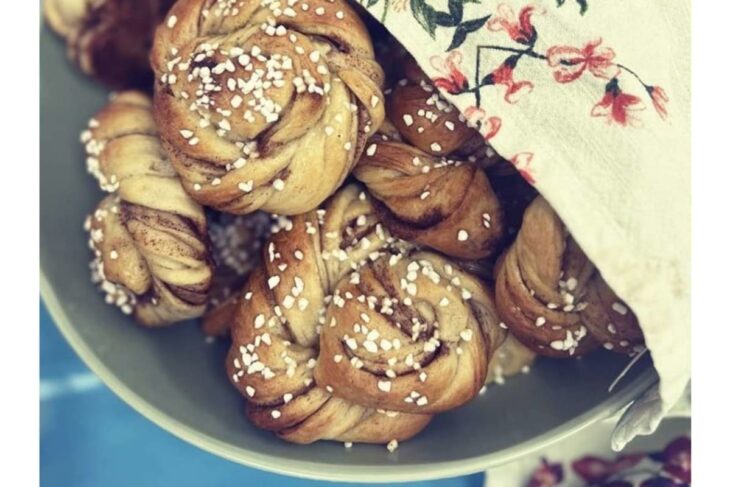Golden brown twisted buns topped with pearl sugar in a ceramic bowl, partially covered by a floral cloth.