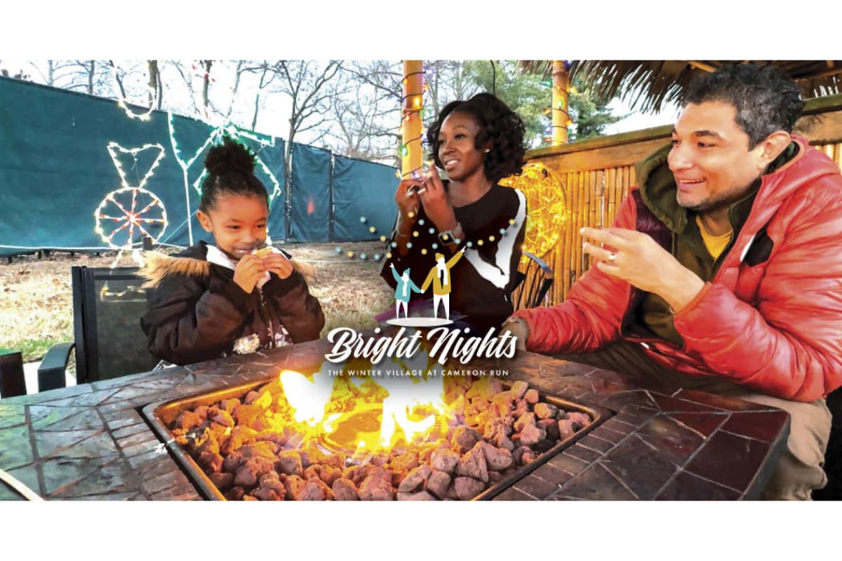 Families enjoy an outdoor fireside moment at Bright Nights: The Winter Village at Cameron Run; a child and two adults smile near a glowing fire pit with festive lights in the background.