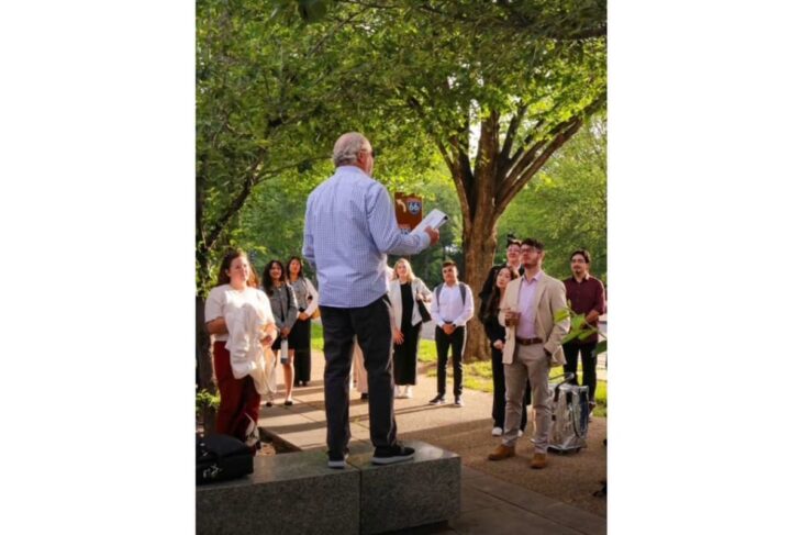 Older man standing on a low platform addressing a small group outdoors under trees.