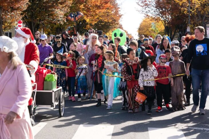 Group of costumed children marching in a Halloween parade on a sunny street, with spectators and a Santa in the foreground.