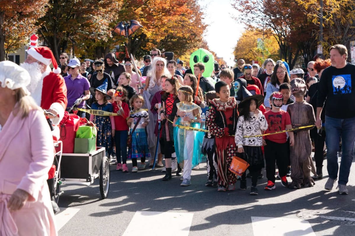 Group of costumed children marching in a Halloween parade on a sunny street, with spectators and a Santa in the foreground.