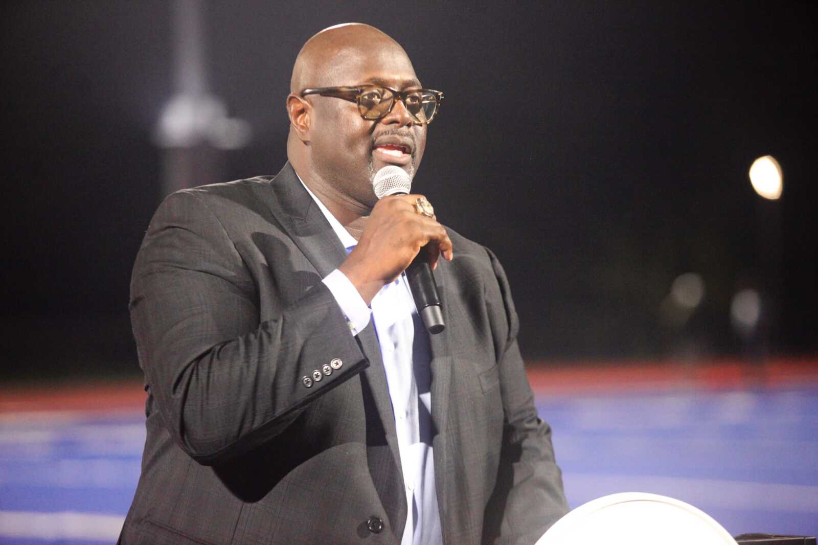 Man in a dark suit and glasses speaking into a handheld microphone on a stage at night.
