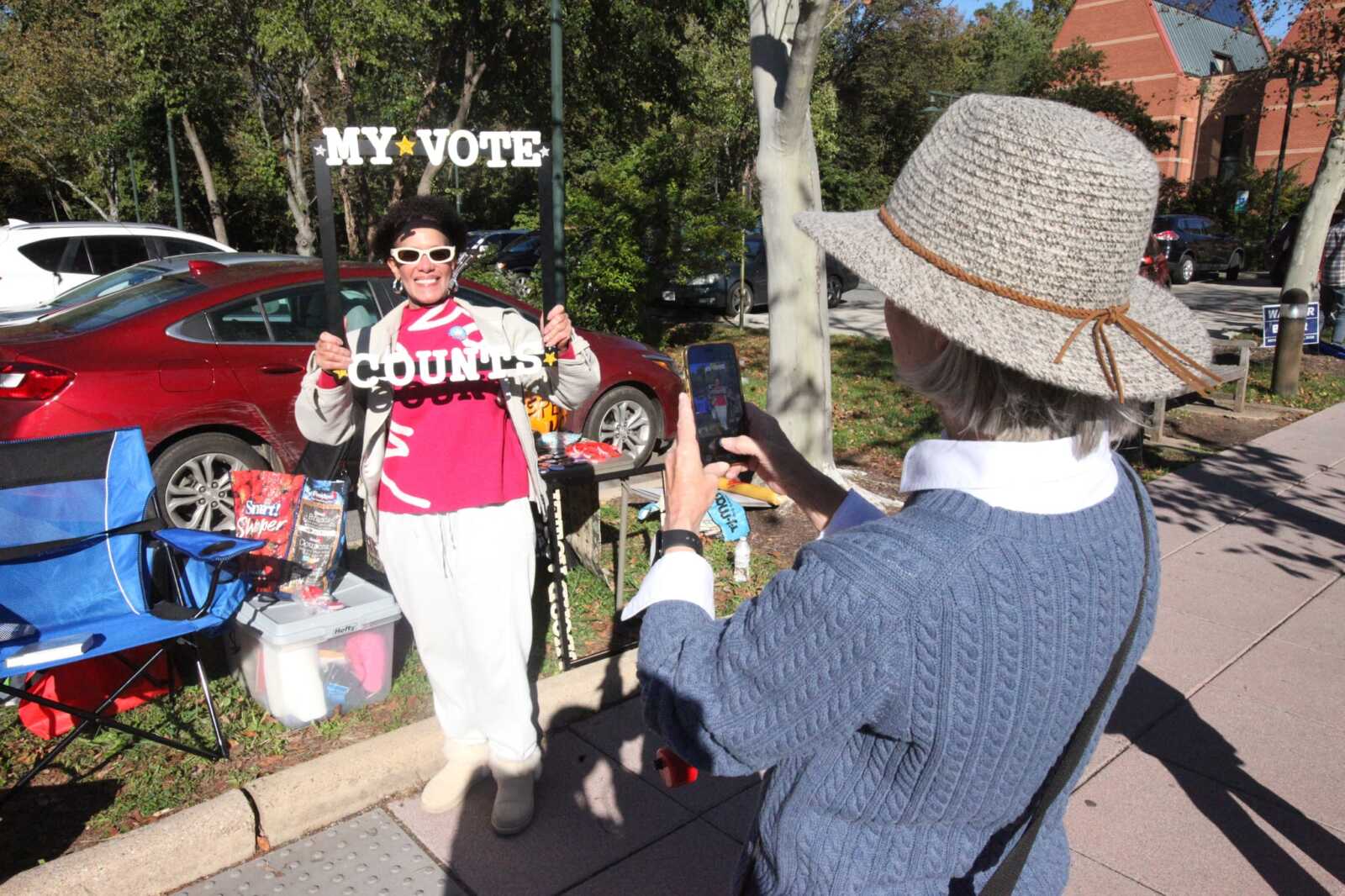 Woman wearing pink shirt holds a 'MY VOTE COUNTS' sign, smiling for a photo outdoors while another person takes her picture in a park setting.