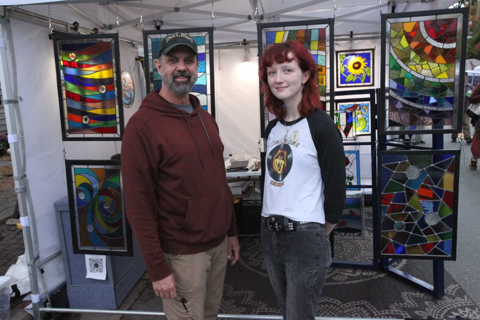 Two people stand beside a display of colorful stained-glass panels at an outdoor market stall.