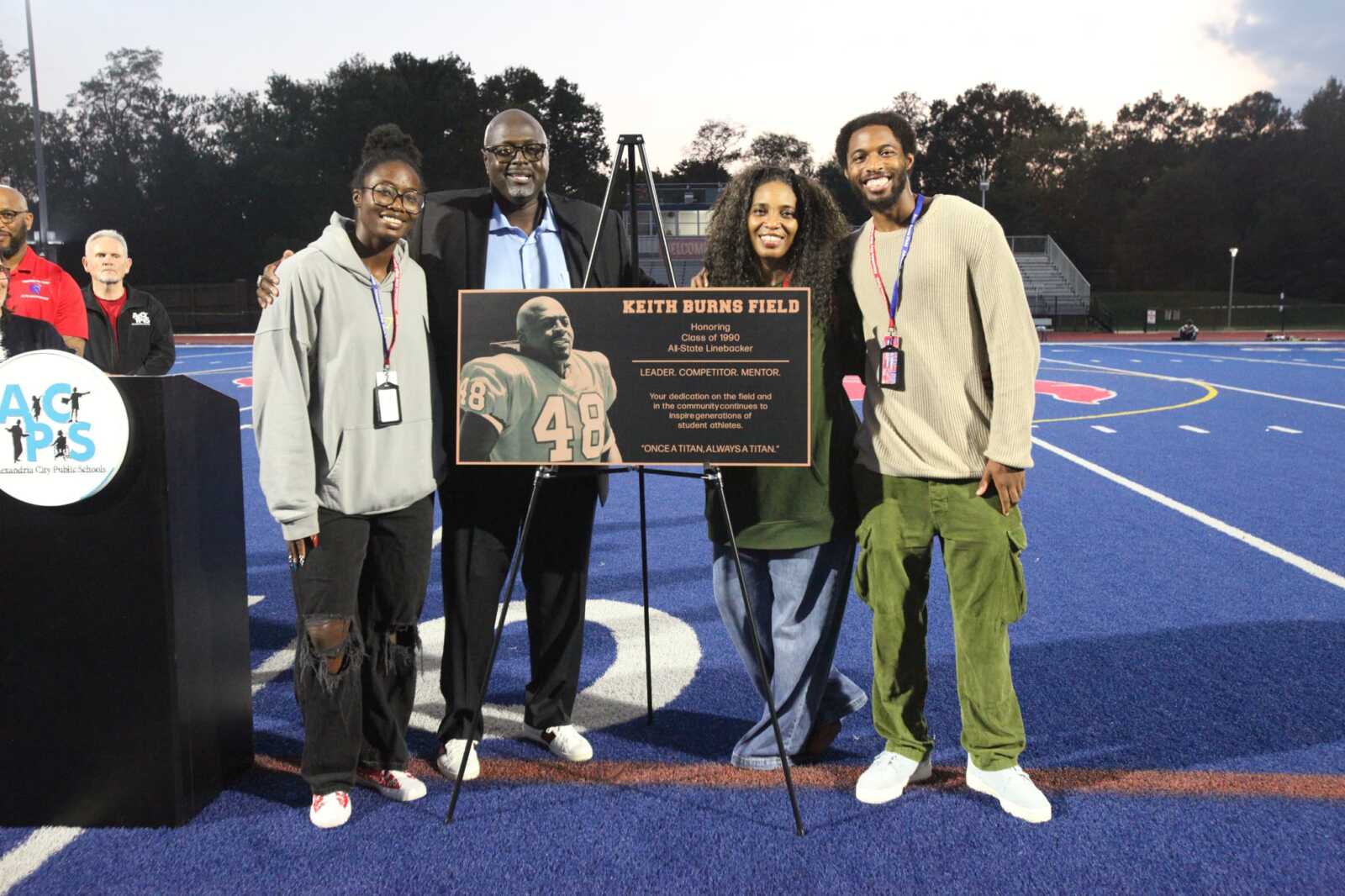 Five people stand on a blue football field beside a commemorative plaque reading Keith Burns Field
